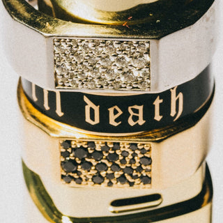 Close-up of stacked rings with a silver band featuring a black enamel section with gold gothic lettering that reads 'in death', flanked by gold-toned rings with textured clear and black stones.