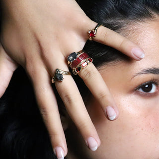 A close-up lifestyle shot of a model's hand wearing multiple stacked rings, including a prominent red and gold ring, against a dark background.