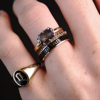 A close-up of a hand wearing multiple rings, including a gold signet ring with a black face and a letter P, a gold ring with a smoky gemstone, and a gold and black band engraved with 'LOVE 24/7'.