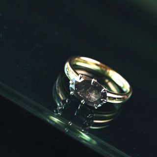A gold ring with a large, faceted dark gray or smoky crystal set in a prong setting, resting on a reflective dark surface.