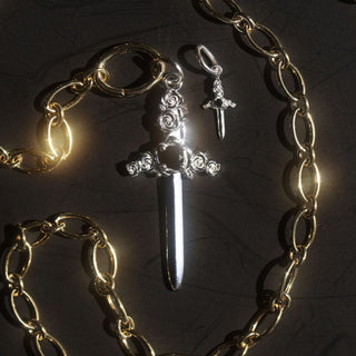 two silver dagger charms (one large and one small) against a black baground with gold chain wrapped around
