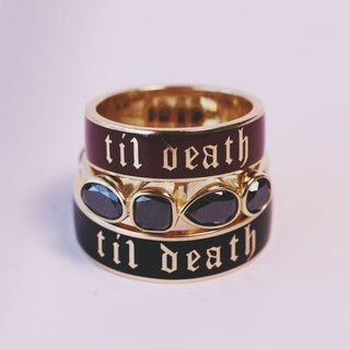 Three stacked gothic-style rings with gold-tone metal and black enamel, featuring the gothic text 'til death' and dark teardrop-shaped stones, set against a soft light purple background.