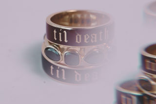 A close-up of a gold ring with a dark enamel band engraved with the gothic-style text 'til death'.
