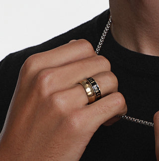 Hand wearing a gold ring with engraved text, against a neutral background