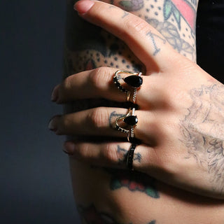 Tattooed hand wearing two black onyx rings and black diamond wave stacking bands, dark background