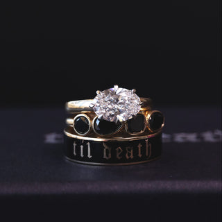 A stack of gothic-style rings on a dark surface against a black background. The top ring features a large round clear gemstone in a gold setting, while the middle ring is black with gold lettering that reads 'til death', and the bottom ring is black with gold circular accents.