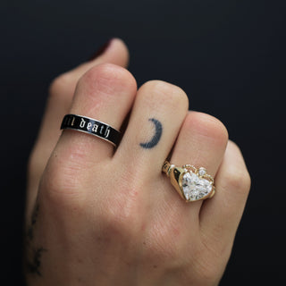 Hand wearing two rings with 'ill death' and heart designs on a dark background
