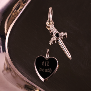 Silver dagger and heart-shaped pendant with 'til death' engraving on a reflective surface.