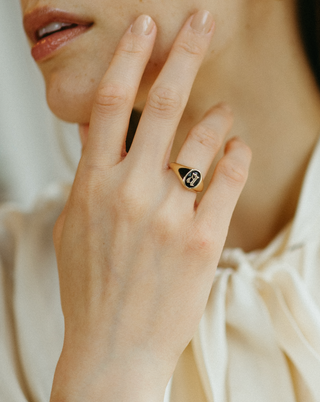Close-up of a woman's hand wearing a gold ring with a black stone, touching her face.