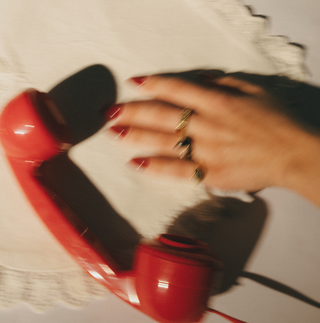 Red phone receiver held by a hand with red nail polish on a light-colored surface.