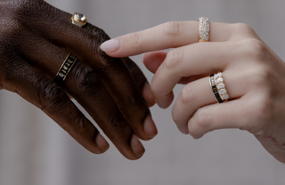 Two hands, one with a dark skin tone and the other with a light skin tone, wearing multiple rings on a neutral background.