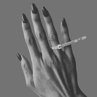 A close-up of a hand with long dark manicured nails wearing a silver ring with a clear rhinestone band, alongside a white plastic ring sizer tool on the middle finger, against a solid gray background.