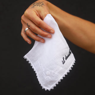 Hand holding a white handkerchief with 'til death' embroidery against a black background