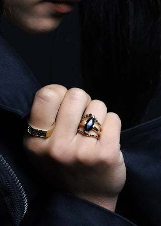 A close-up lifestyle shot of a model wearing a gold ring with a black oval stone on her finger, paired with a black jacket against a dark background.