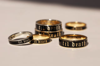 Several black and gold rings engraved with the gothic-style text 'til death' arranged on a light neutral surface.