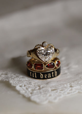 Stack of three rings against a neutral beige background: a heart-shaped diamond engagement ring, a mixed shape garnet band, and a til death enamel black ring - all in yellow gold.