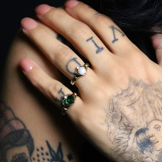 Tattooed hand wearing the Opal Sempre and Tsavorite Sempre heart rings on a dark background