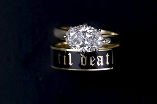 A gold and black ring set featuring a round-cut diamond solitaire ring on top of a black band engraved with the gothic-style text 'til death'.