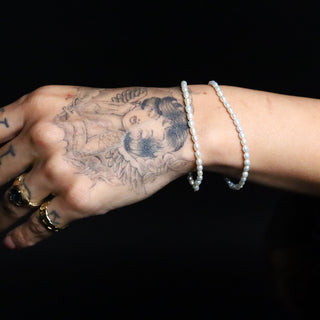 Two Everyday Tear Drop Pearl Bracelets on a tattooed wrist with gold rings, against a black background