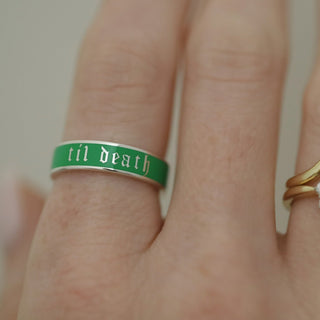 Hand wearing three rings with 'til death' on a green band, gold ring with diamonds, and gold stacking band.