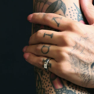 Tattooed forearm with floral tattoos and a sterling silver Til Death band on the finger, dark background