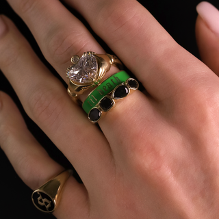Close-up of a hand wearing a gold ring with a large diamond and green stone on a black background