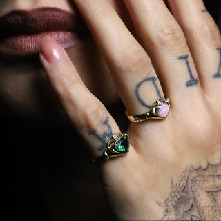 Tattooed hand wearing the green Tsavorite Sempre Ring and a second heart Sempre ring on a dark background